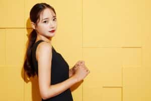 黄色い背景の前に横に立ってこちらを見る女性